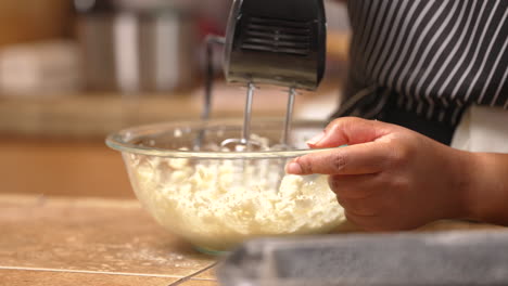 side view of a black woman whipping butter for batter with a hand mixer in a glass bowl