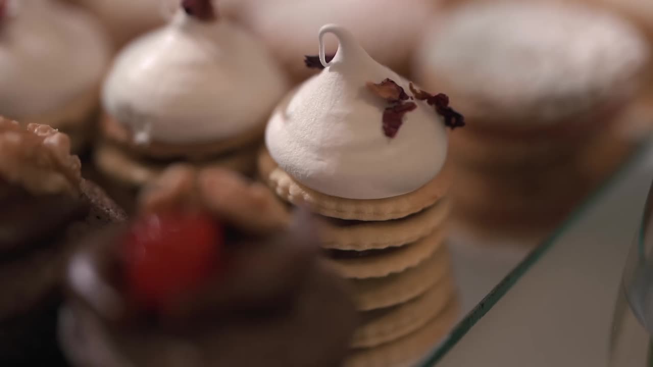 Close-up panning shot of assorted mini pastries topped with meringue, raspberries, nuts, chocolate, and edible petals, arranged on a wedding sweet table with shallow depth of field