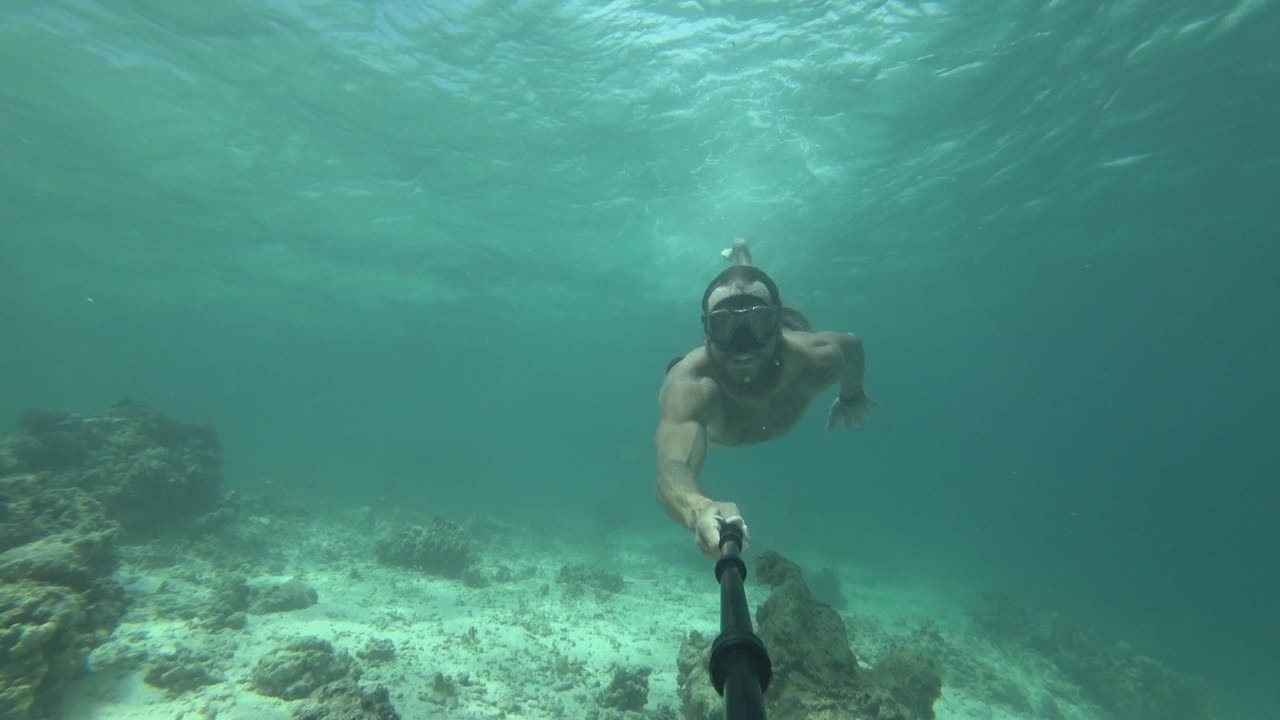 un hombre joven, en forma y fuerte con pelo largo y barba está nadando con gafas de esnórquel y buceando desde la superficie hacia el agua vibrante y va entre un espacio estrecho entre el arrecife