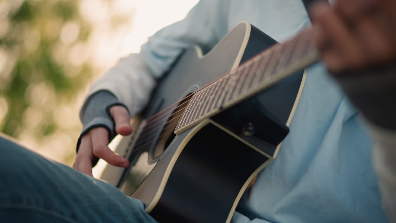 artista di strada bianco che suona la chitarra acustica su una panchina, primi piani a livello della vita, ambientazione rilassata in un parco, jeans di denim, ritmo costante, controluce naturale, atmosfera spontanea da esibizione di strada, luce solare calda