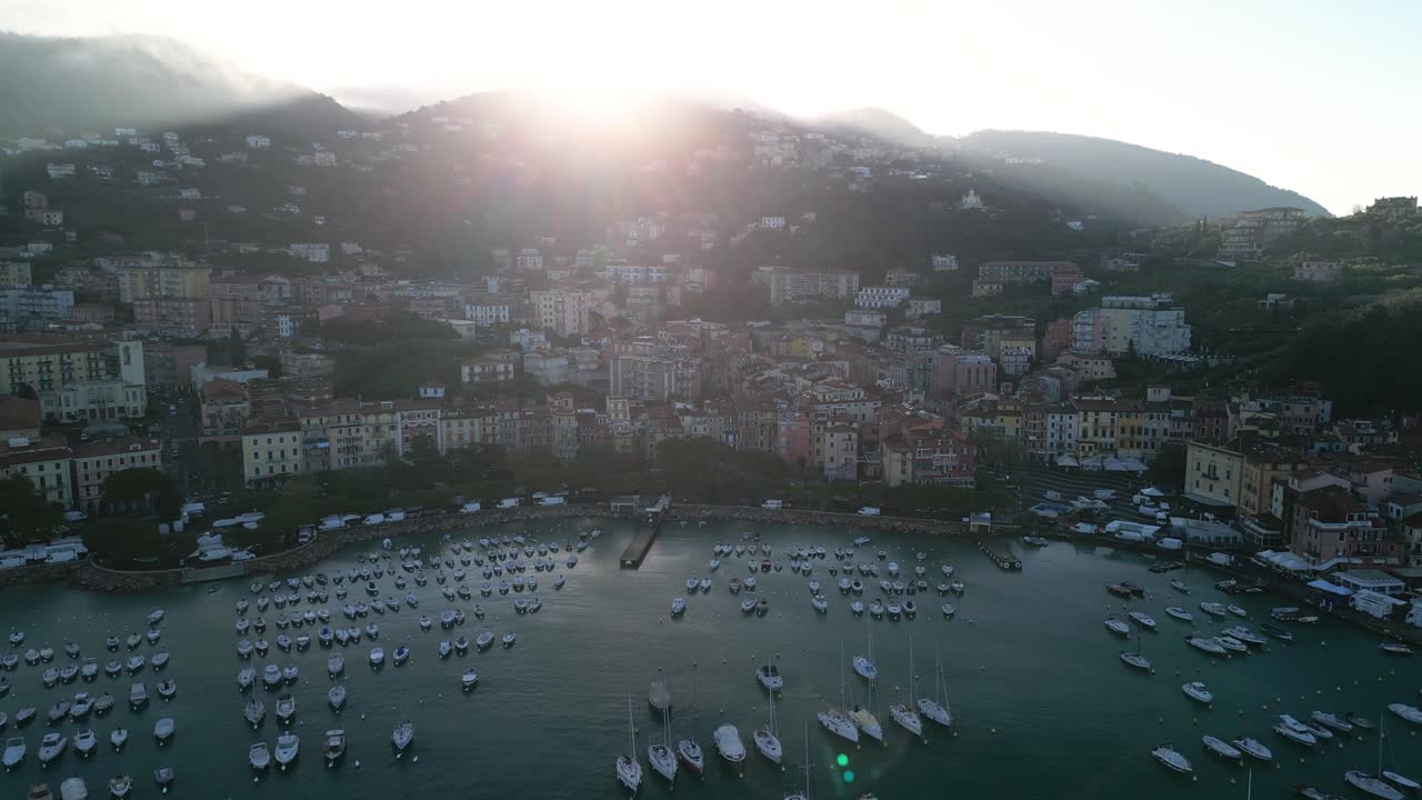 Ripresa aerea con drone in allontanamento sopra le barche nel porto turistico di Lerici, Italia, mentre l'alba si fa strada attraverso la nebbia sulla cima della montagna che fa da sfondo al paese di mare.