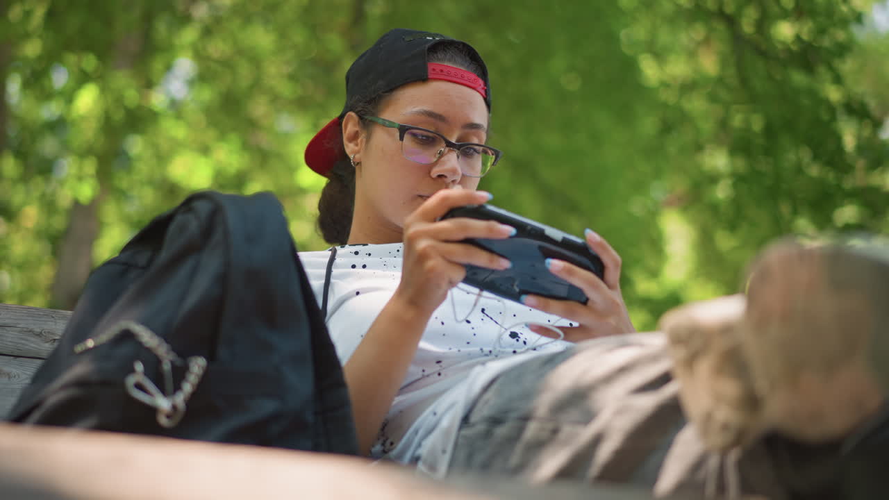 adolescente jugando con una consola portátil en un banco; joven con gafas y gorra al revés desplazándose por un juego con expresión concentrada; mochila a un lado; luz solar moteada entre los árboles; pulgares tecleando los controles