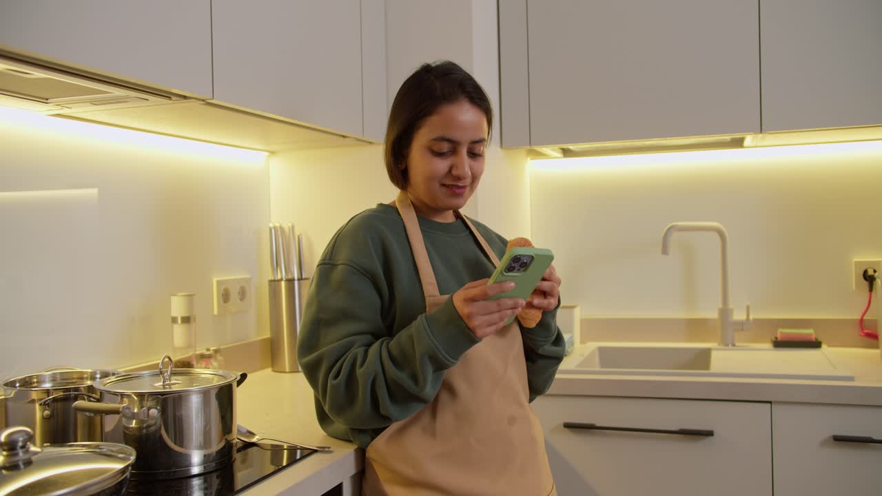 una chica india morena feliz con un suéter verde y un delantal beige come un croissant y escribe en su teléfono inteligente verde en la red social durante su descanso y almuerzo en casa en un apartamento moderno