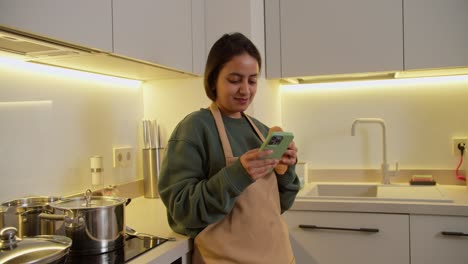 Una-Chica-India-Morena-Feliz-Con-Un-Suéter-Verde-Y-Un-Delantal-Beige-Come-Un-Croissant-Y-Escribe-En-Su-Teléfono-Inteligente-Verde-En-La-Red-Social-Durante-Su-Descanso-Y-Almuerzo-En-Casa-En-Un-Apartamento-Moderno