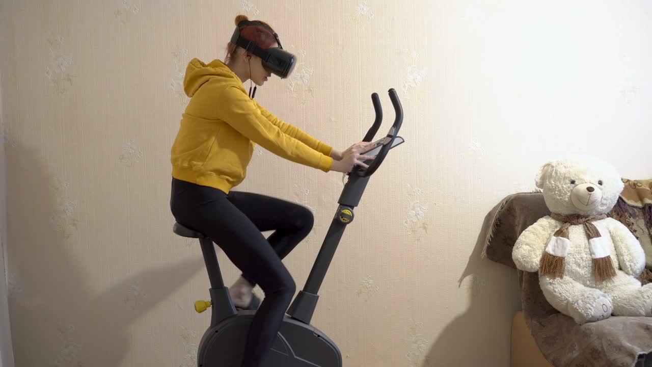 어린 소녀가 가정에서 운동 자전거를 타고 가상 현실 안경을 착용하고 헤드폰을 통해 음악을 듣고, 노란색 스포츠 옷을 입은 소녀, 가상 훈련의 개념.