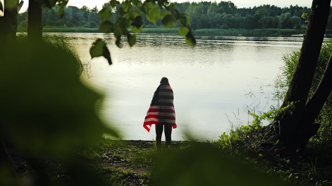 Patriotic woman draped in USA flag watches sunset in slow motion forest view