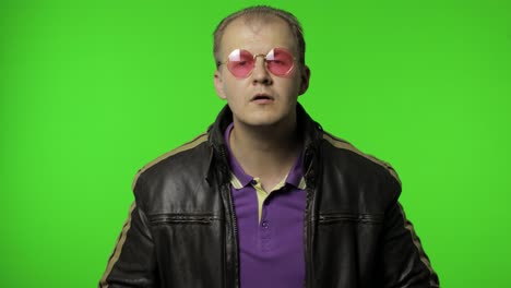 cool brutal rocker man in brown denim jacket puts on pink sunglasses, looks at camera and smiles