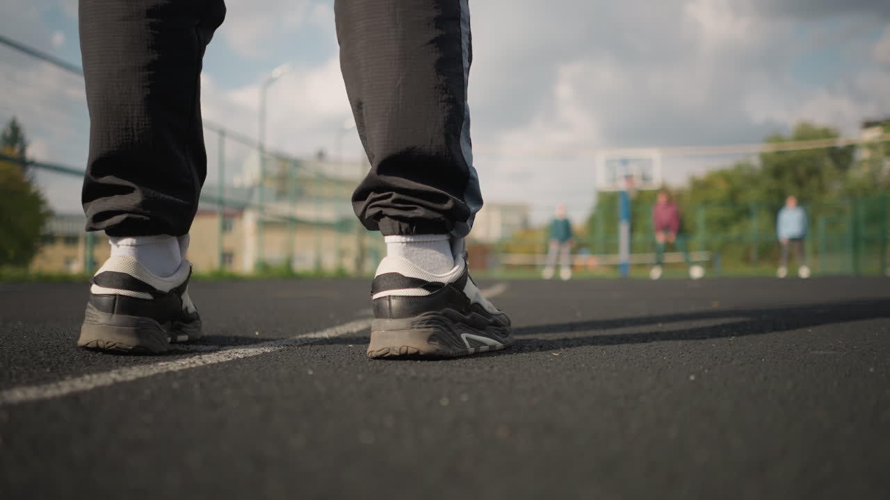 vista trasera de un atleta con zapatillas negras rebotando la pelota de voleibol en la cancha con figuras borrosas de tres personas en el fondo, rodeadas de vegetación