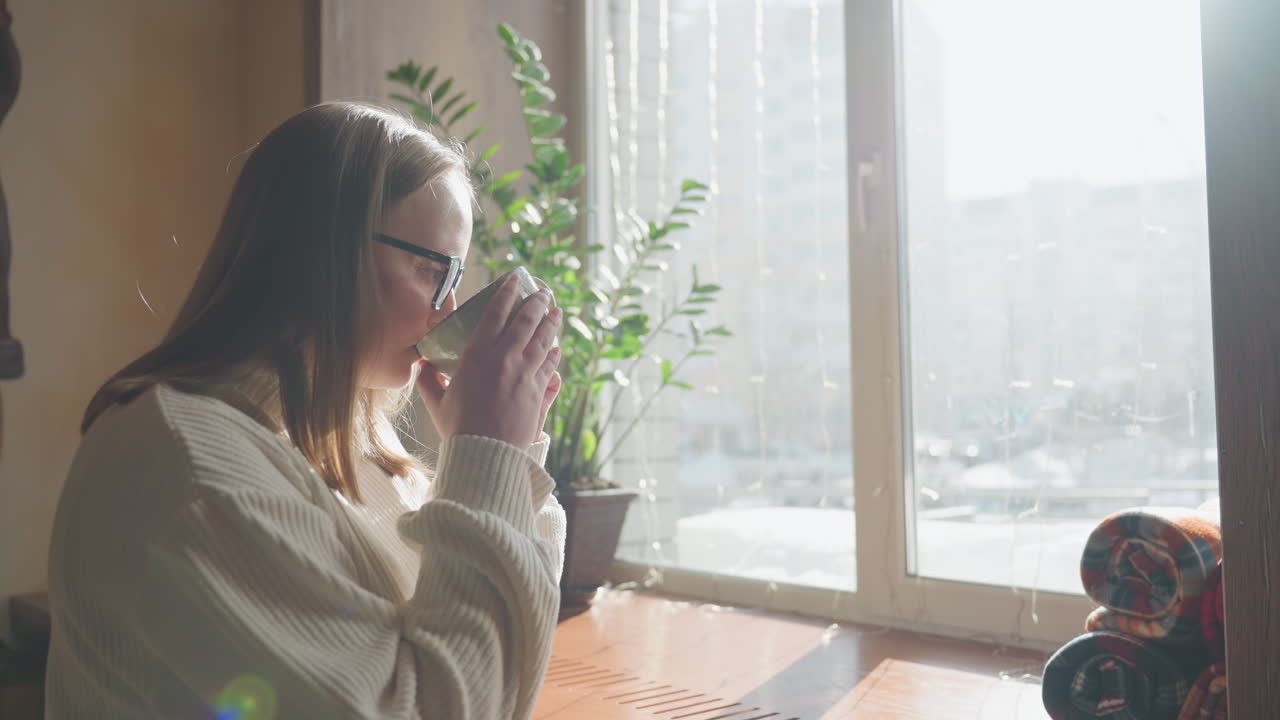Woman sits by window with warm sunlight pouring in, holding coffee cup near potted plant, calm indoor setting with natural light, cozy ambiance, book on table, and mural partially visible on wall