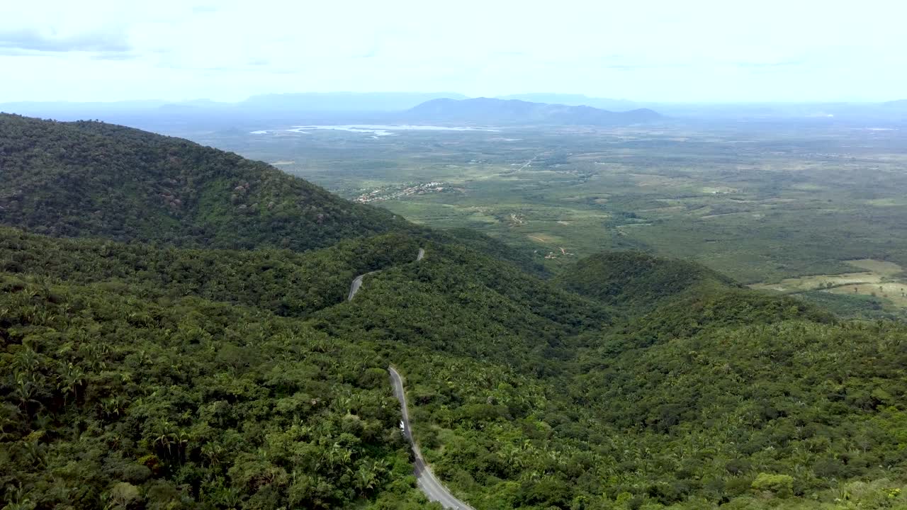 Aerial drone shot shows winding road cutting through lush green forested hills with wide valley and distant mountains in background, showcasing remote natural beauty of Canadian landscape