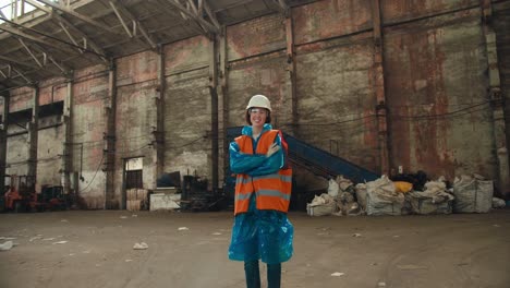 Close-up-portrait-of-a-brunette-girl-in-a-blue-protective-uniform-and-an-orange-vest-who-stands-in-the-huge-hall-of-a-waste-processing-plant-and-smiles