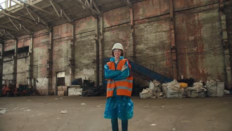 retrato en primer plano de una chica morena en un uniforme de protección azul y un chaleco naranja que se encuentra en el enorme salón de una planta de procesamiento de residuos y sonríe