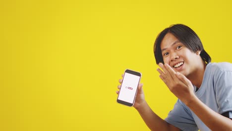 Young-Asian-Male-Student-Pointing-At-Smartphone-Screen-With-Like-Button-And-Showing-Inviting-Gesture