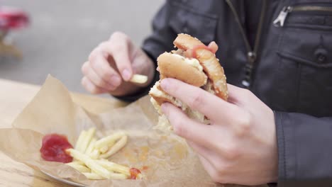 young man with beard in the street cafe eating french fries and biting tasty big burger with cheese. shot in 4k