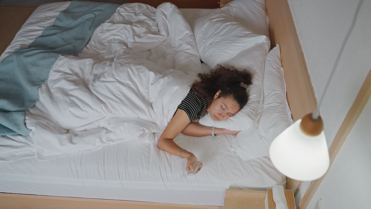Fredfyldt morgensøvn. Fredelig opvågning under bløde hvide lagner. Blid morgenstemning med hvilende kvinde og varmt lys. Afslappende tidlig morgenstemning med kvinde hvilende i et veloplyst soveværelse.