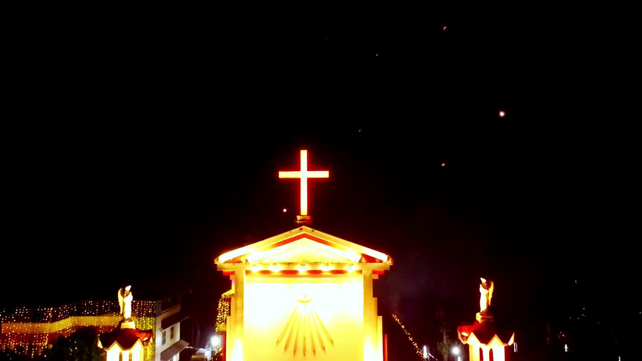 夜間、明るくライトアップされた教会のドローン空撮。輝く赤い十字架と鮮やかな色彩に変化するLEDライトが特徴で、暗い夜空には祝祭の花火が打ち上がっている。