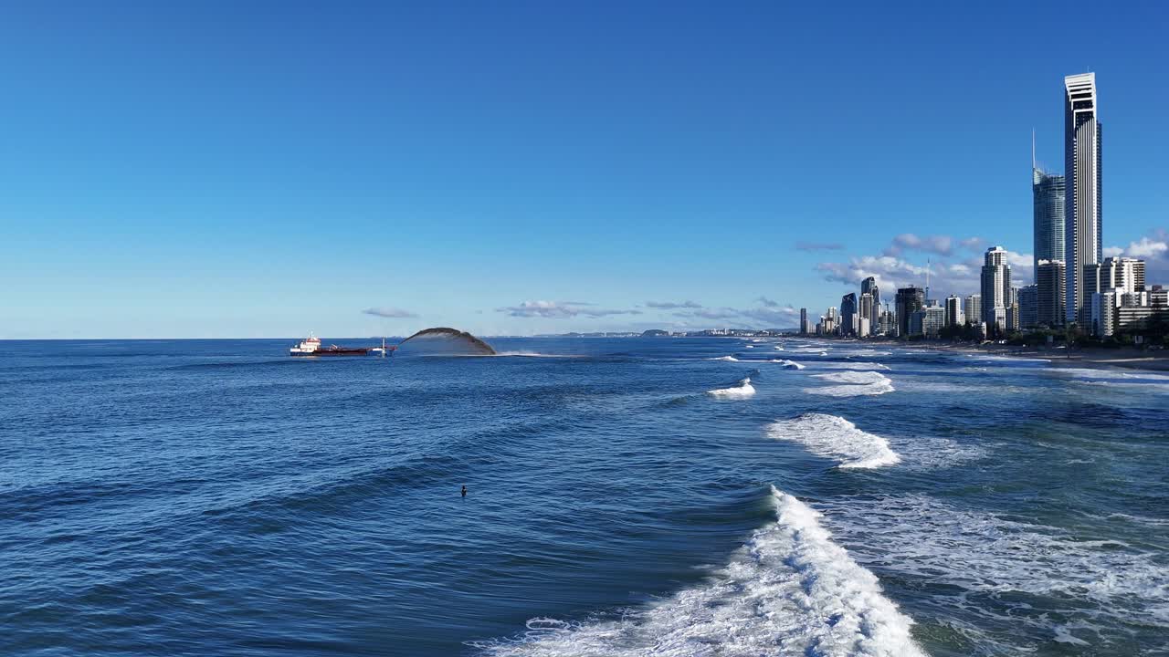 A dredger pumps sand in the Pacific Ocean near Gold Coast's skyline, under clear blue skies with gentle waves