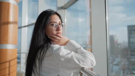 stylish young woman in white shirt and glasses rests on railing, gazing thoughtfully outside through large glass wall with cityscape in background under soft natural daylight