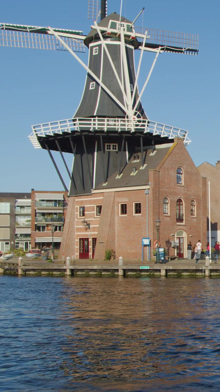 Motoscafo con passeggeri passa accanto a un mulino a vento olandese storico sul canale di Haarlem sotto un cielo limpido e diurno