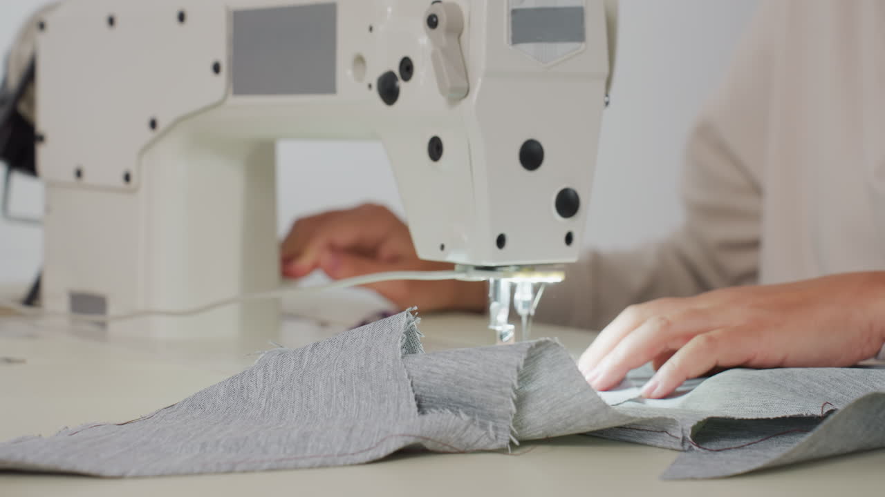 Seamstress sewing dress on industrial machine with bright light, focusing on precise stitching, controlled tension, and skillful approach to transform gray fabric into garment in minimal workspace