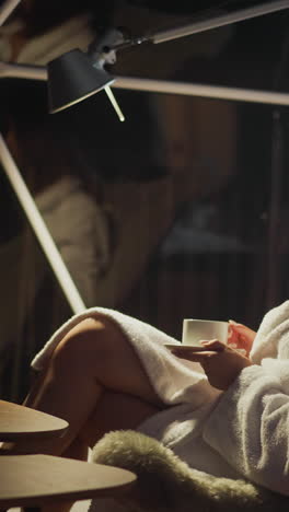 balanced woman resting lit by light of lantern enjoys evening coffee after working day. young woman sitting in armchair calmly looks away in romantic atmosphere