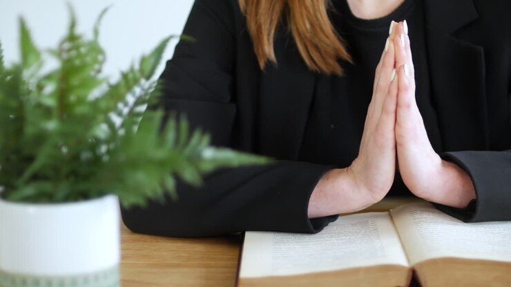 A woman praying with hands held flat together on an open bible book on a table