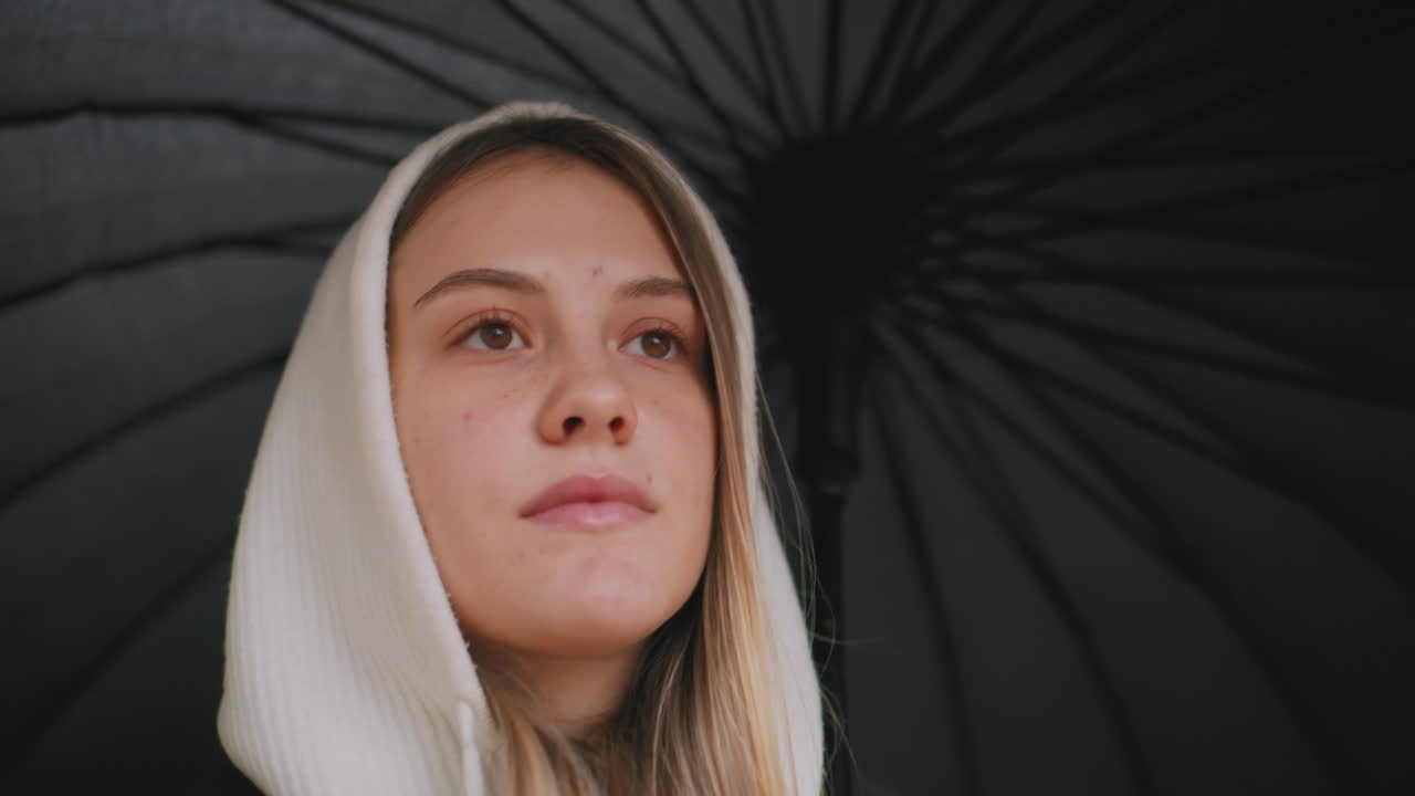 Closeup portrait of young woman in white hood under black umbrella, looking forward with calm expression on face, outdoor autumn rainy weather, thoughtful mood, peaceful vibe, natural light detail