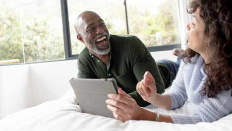 happy mature diverse couple lying on bed and using tablet at home, slow motion