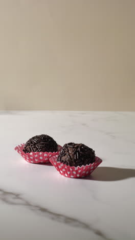 close-up of a hand placing a chocolate truffle with sprinkles into a red polka dot paper cup on a marble surface. bright lighting and minimal background highlight the elegant, handmade dessert.