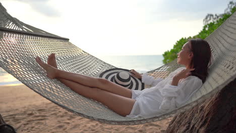 young barefoot beautiful thai woman with large black and white hat swings on hammock by the sea