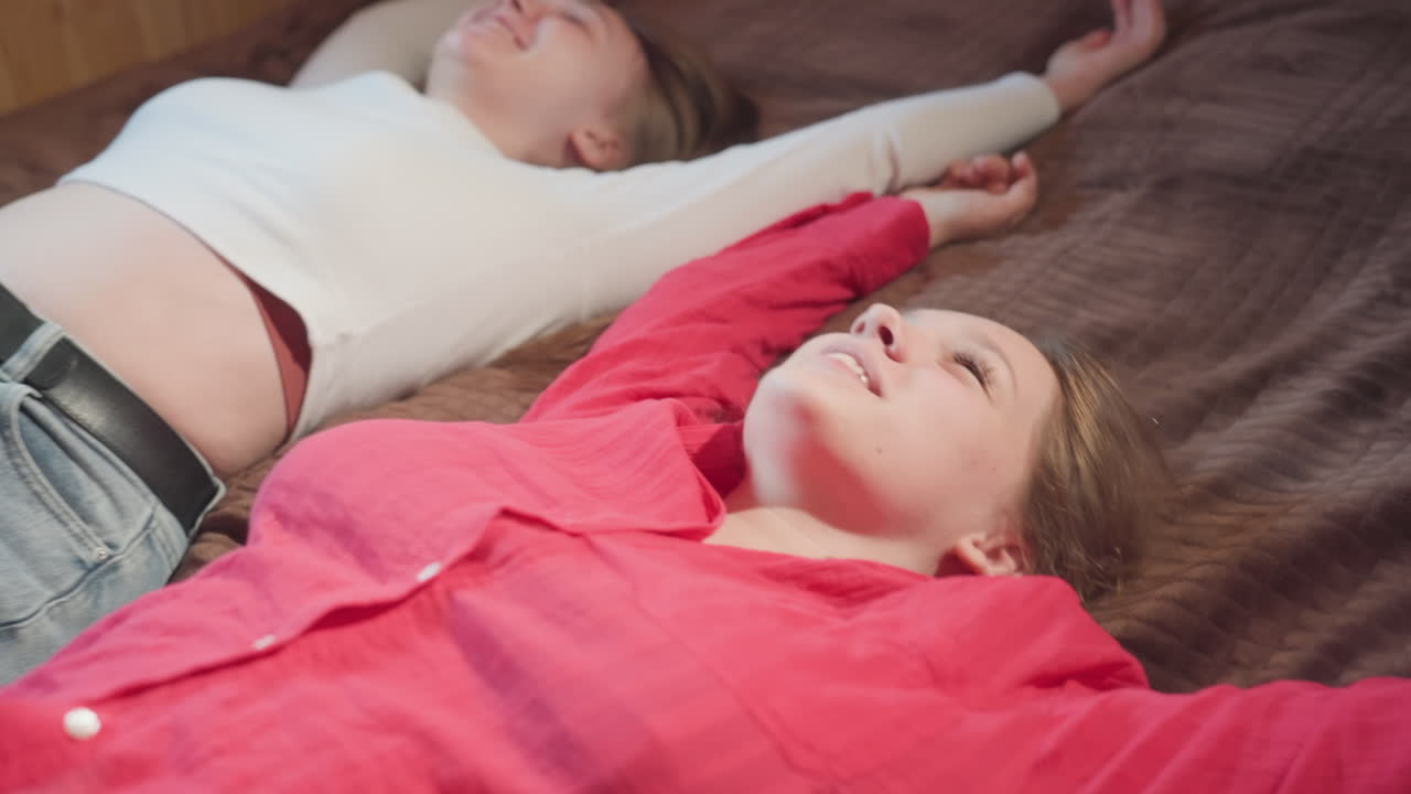 close up of joyful young women falling backward onto bed in warmly lit wooden room, smiling and enjoying carefree playful moment together while sunlight streams through window in cozy setting