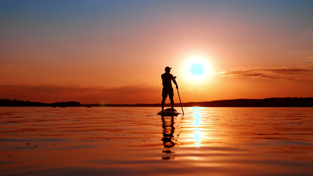 Male silhouette distancing from camera on the paddle board. Man oars standing on the inflatable sup board floating by the river at sunset. Low angle view.
