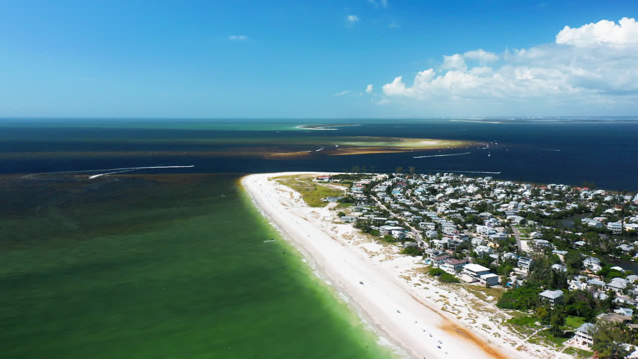 A coastal peninsula lined with homes extends along a sandy beach where emerald shallows meet offshore deep waters, capturing the meeting point of community and open sea