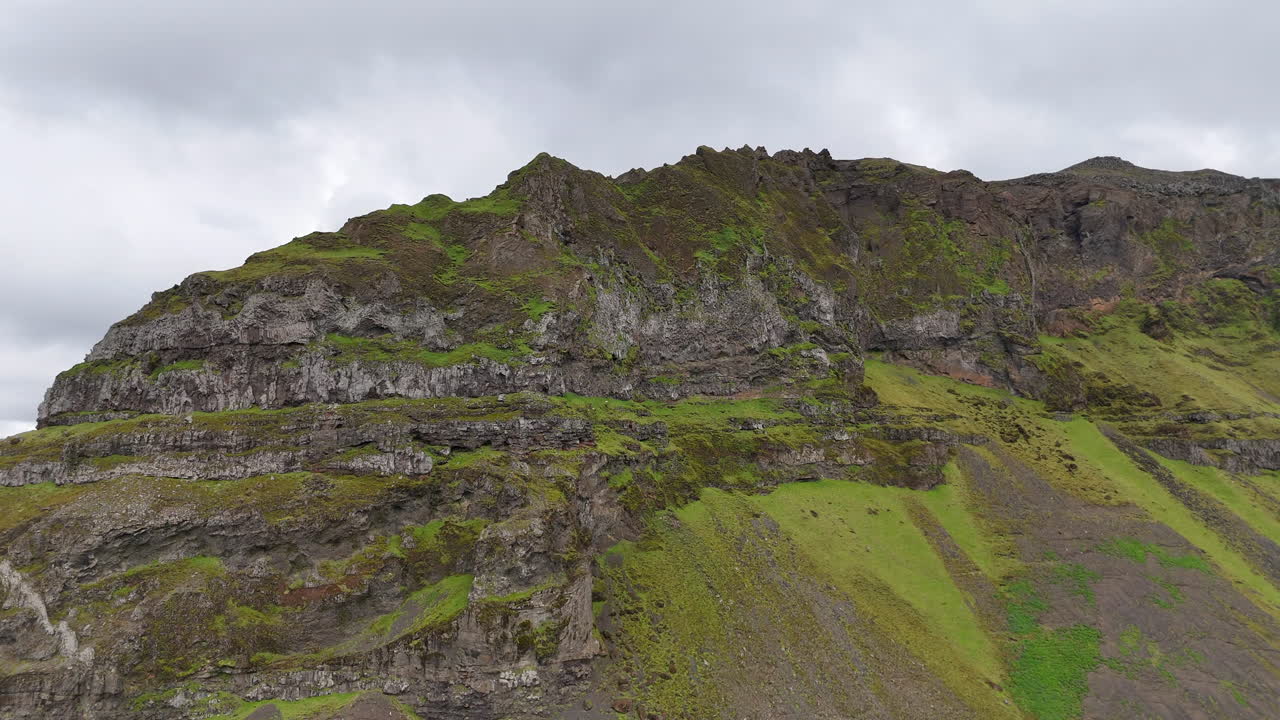 Aerial view of vibrant green fields stretching below steep moss-covered cliffs in Suðurland, Iceland, with winding streams and dramatic shadows shaping the vast volcanic landscape