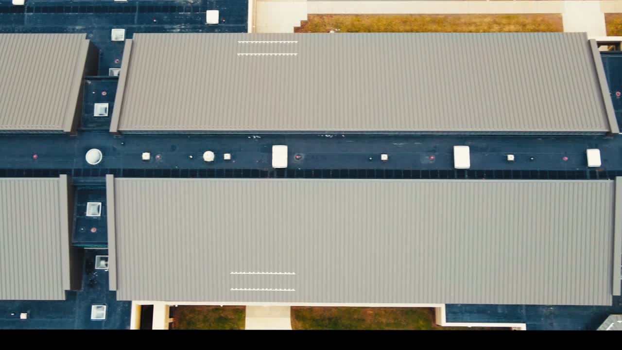 A precise top-down drone view of a large modern school roof with clean metal panels and multiple ventilation units. The symmetrical roof design is surrounded by well-maintained green spaces.