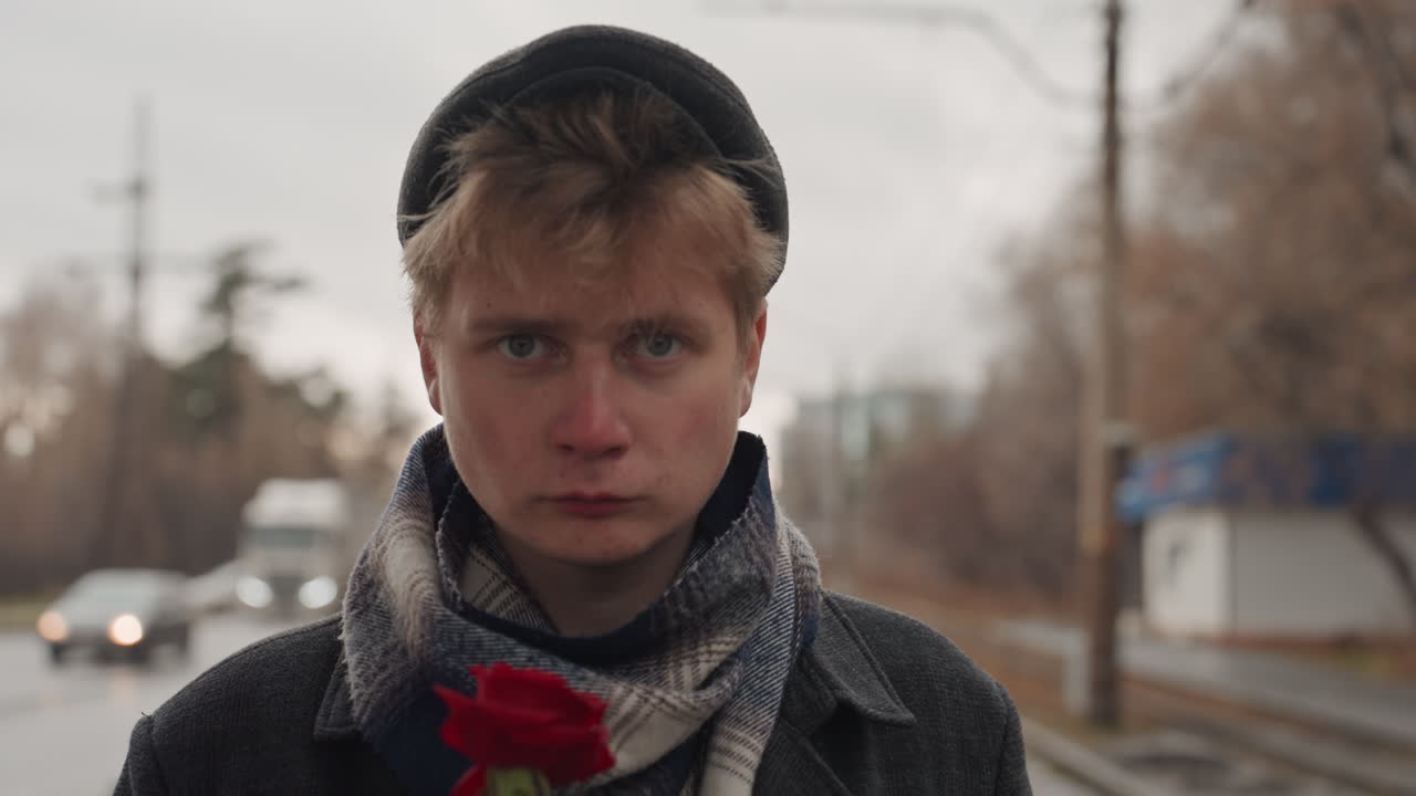 Young man on rainy city sidewalk with rose. Closeup of pensive man holding red rose in rainy city atmosphere. Contemplative young man stands on rainy urban street holding vibrant red flower