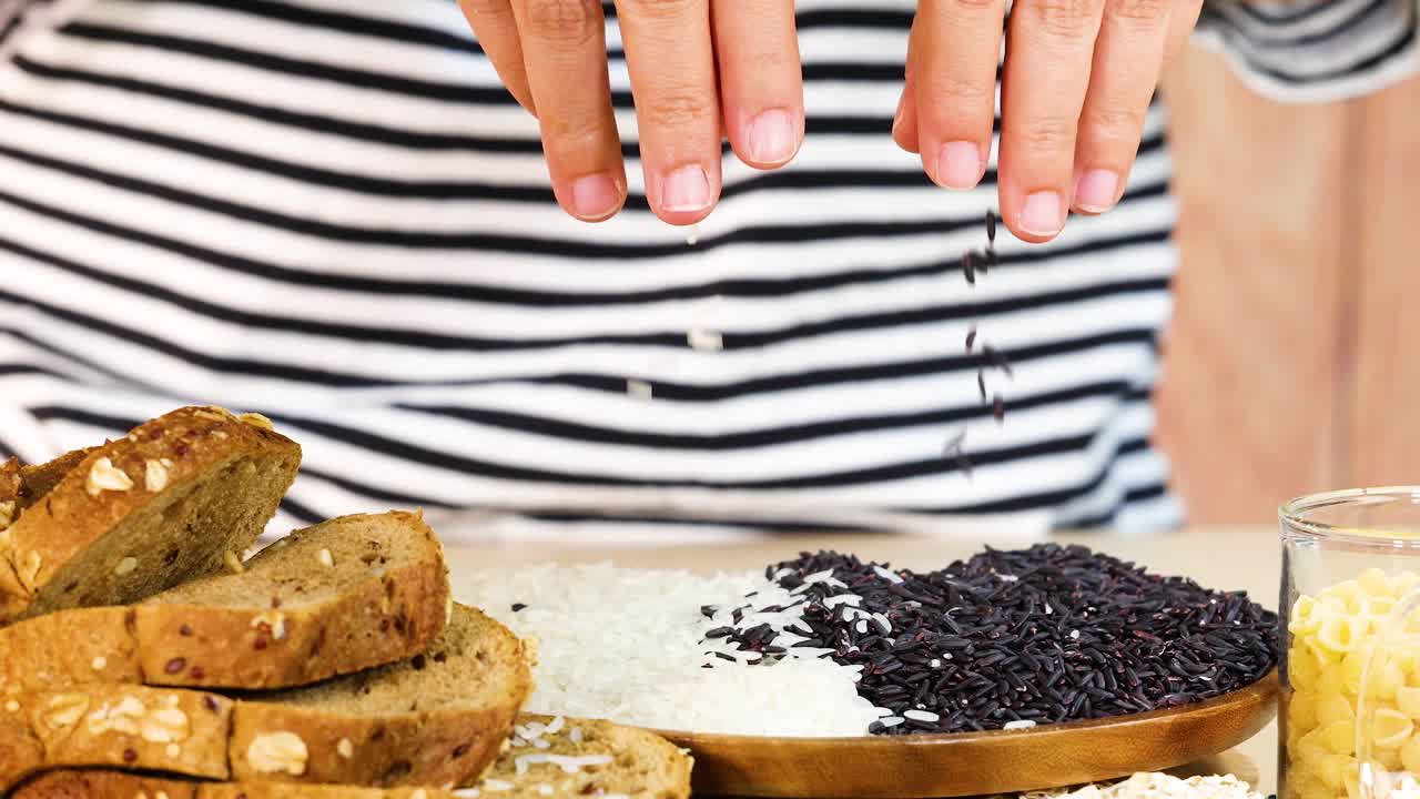 Hands organize various grains and bread slices on a wooden board. Bright lighting highlights the textures and colors