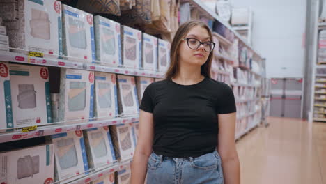 mujer caminando cerca de un estante de supermercado lleno de productos textiles, centrándose en las mercancías exhibidas mientras un grupo de personas pasa en el fondo, entorno minorista con ropa de cama y textiles para el hogar