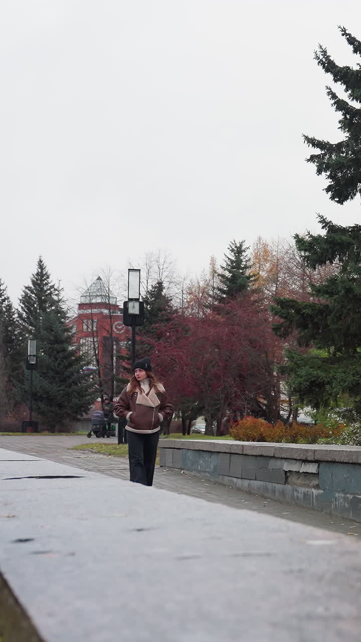 Thoughtful girl wearing black cap brown jacket and trouser walking slowly with hands in pockets on a pathway surrounded by pine trees flower beds modern buildings and overcast sky during cold autumn
