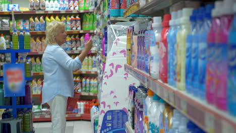 Seniorin-Schlendert-Mit-Einkaufskorb-In-Der-Hand-Durch-Den-Supermarkt,-Dann-Sucht-Sie-Sich-Etwas-Zum-Waschen-Aus-Und-Legt-Es-In-Einen-Einkaufswagen