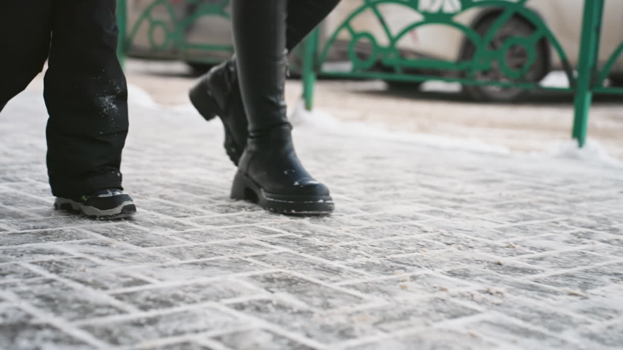 Mamma e figlio bianchi su una strada di ciottoli ghiacciata. La mamma pendolare cammina con determinazione mentre il bambino la segue, indossando un cappotto caldo e stivali da neve. La pavimentazione strutturata è spolverata di neve, con auto parcheggiate e una ringhiera verde.
