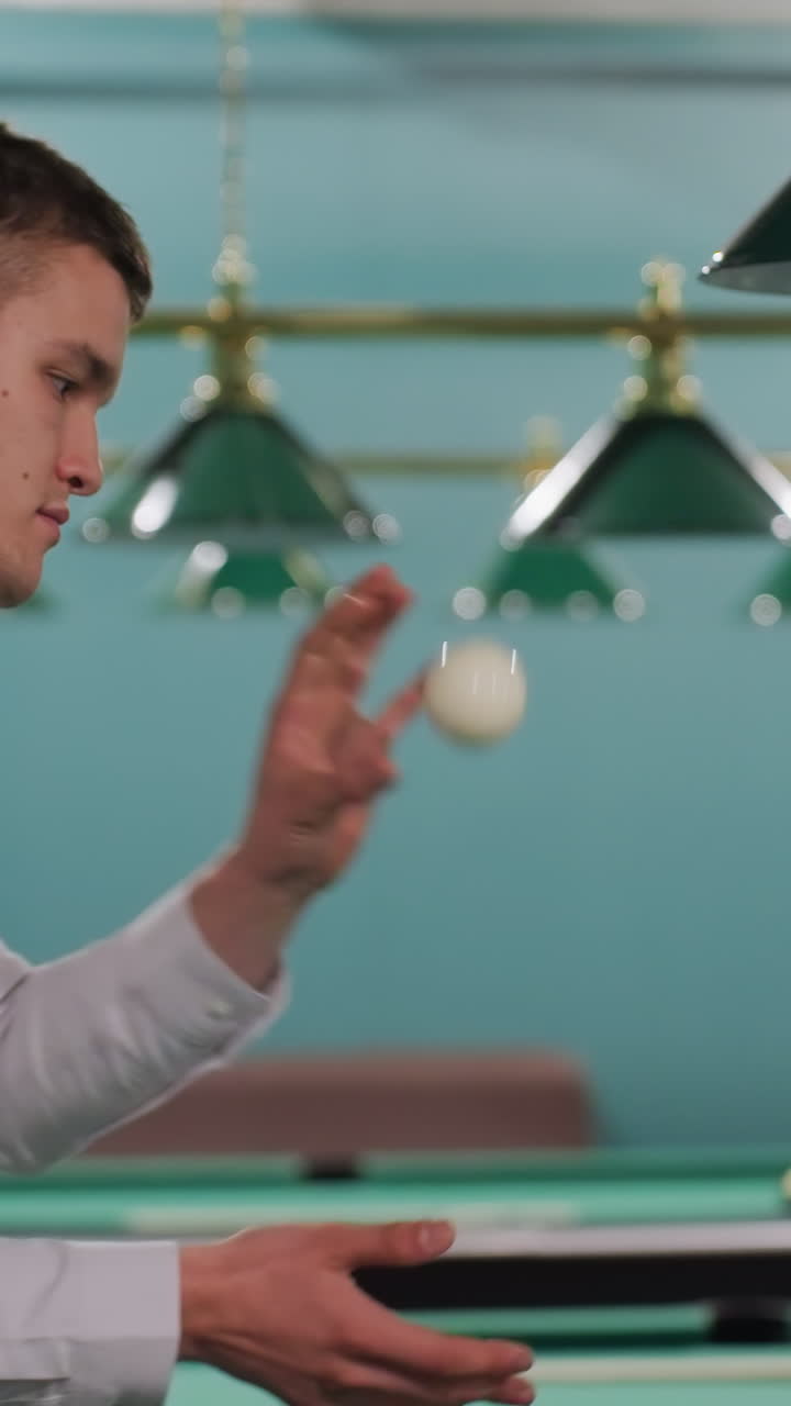 caballero con camisa blanca caminando en la sala de billar mientras recoge la pelota de billar blanca y la lanza en el aire. expresión enfocada, atmósfera de club deportivo moderno, mesas de billar verdes