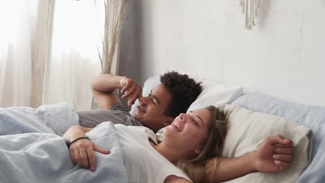 Happy-multiracial-couple-lying-in-bed-and-laughing.-Waking-up-together-in-the-morning.-Attractive-young-man-and-woman-looking-at