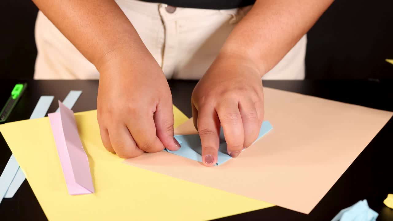 Person folding blue origami paper on yellow sheet, well-lit tabletop, close-up, steady camera