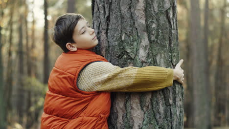 side view of a cute little teen boy hugging a tree trunk with eyes closed in the forest