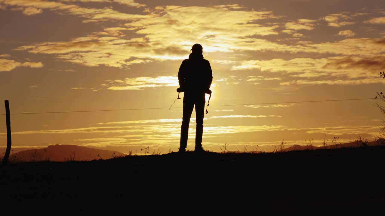 Static wide shot with a man with a backpack walks across the screen, then stops to face the golden sun, standing silhouetted against a dramatic sky. Static wide shot