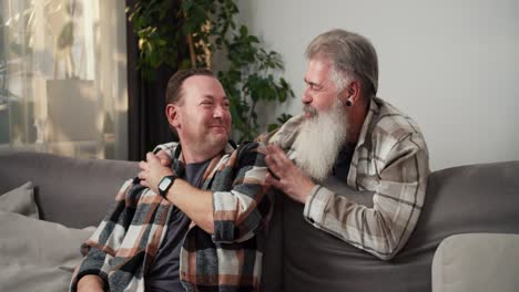 happy elderly man with gray hair and a lush beard in a checkered shirt communicates with his brunette boyfriend and holds hands with him while hanging out at home near the sofa in a modern apartment