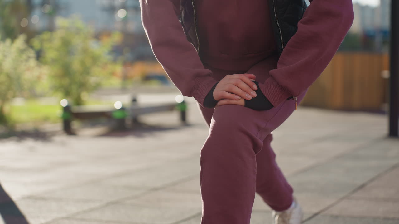Gym instructor hoodie quilted vest performs single leg pistol squat with hands braced on knee under bright sun casting sharp shadows across urban park tiles with blooming flowers modern fitness bars