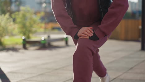 gym instructor hoodie quilted vest performs single leg pistol squat with hands braced on knee under bright sun casting sharp shadows across urban park tiles with blooming flowers modern fitness bars