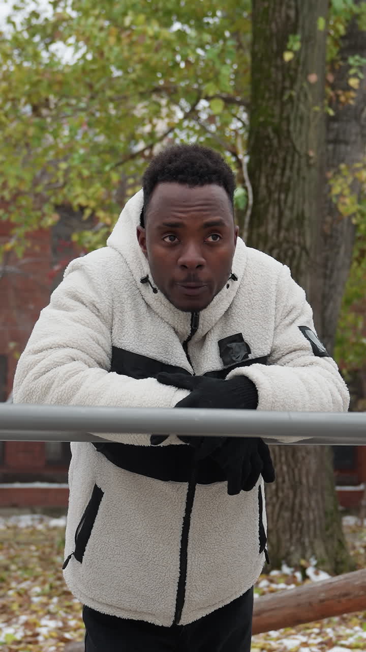Contemplative young adult leans on iron bar, looking around in deep thought, standing in outdoor workout area with snow-covered ground, residential buildings and trees in background